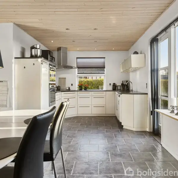 Et moderne køkken med hvide skabe og sorte bordplader, hvor friske blomster står på bordet. Store vinduer lader naturligt lys ind og udsigten viser en have.