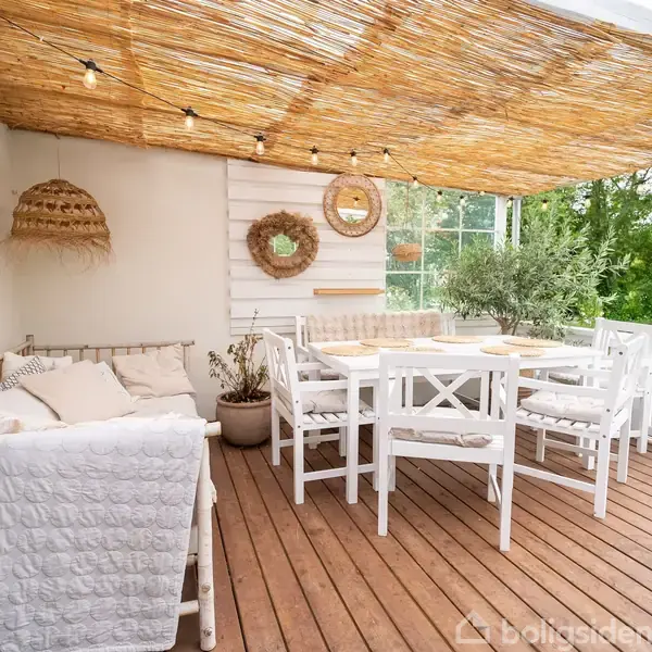 Øverste træterrasse med hvidt møbelsæt, hvor stole og bord står under et bambusloft med lyskæde. Puder og planter skaber en hyggelig atmosfære i en haveomgivelser.