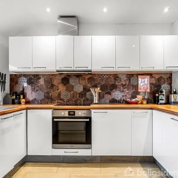 Et hvidt køkken med træbordplade og brun mosaikflisevæg. Der er indbyggede ovn og opvaskemaskine, køkkenredskaber på bordet, og et vindue til venstre giver naturligt lysindfald.