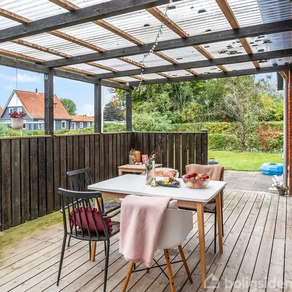 En overdækket terrasse med et træbord og stole er pyntet med mad og drikkevarer. Bagved er en have med græs og træer. Rødt murstenshus ses til højre. Ingen tekst til stede.