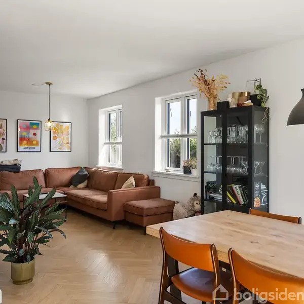 En stue med en brun hjørnesofa står mod væggen under malerier. Ved siden af sofaen står et vitrineskab med glas. Et spisebord med stole er i forgrunden. En plante står til venstre. En ur hænger på væggen.
