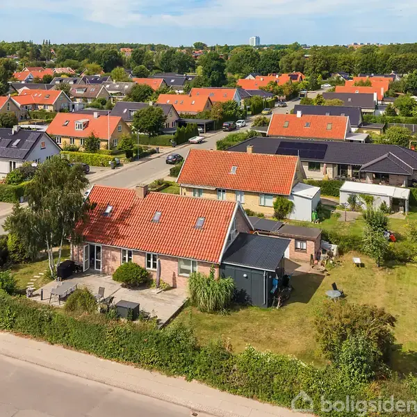 Røde huse med tegltage står i en række i en forstad. En grusvej omgiver dem, og frodige haver adskiller boligerne. En bil er parkeret på vejen.