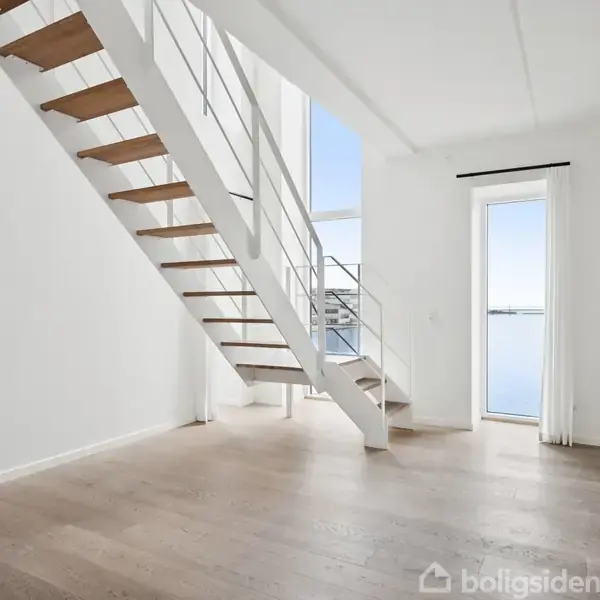 Trappe fører opad i en lys stue med trægulv. Store glaspartier giver udsigt til himmel og vand, mens lyset strømmer ind. Rommet er moderne og minimalistisk.