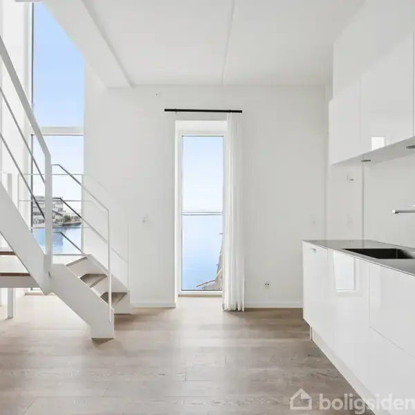 En moderne trappe af træ og metal fører til en øvre etage, mens et lyst køkken med hvide skabe og sort vask er placeret til højre. Store vinduer viser havudsigt.