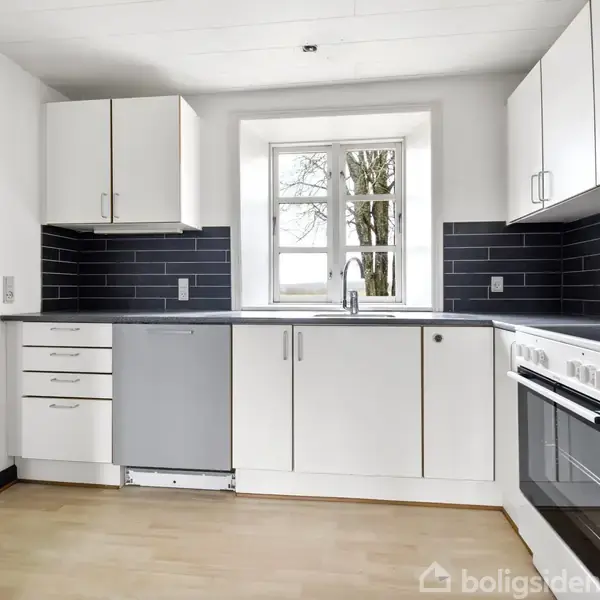 Køkken med hvide skabe, grå opvaskemaskine og ovn, sort flisebagvæg, trælaminatgulv. Et vindue centralt over vasken viser et træ udenfor.