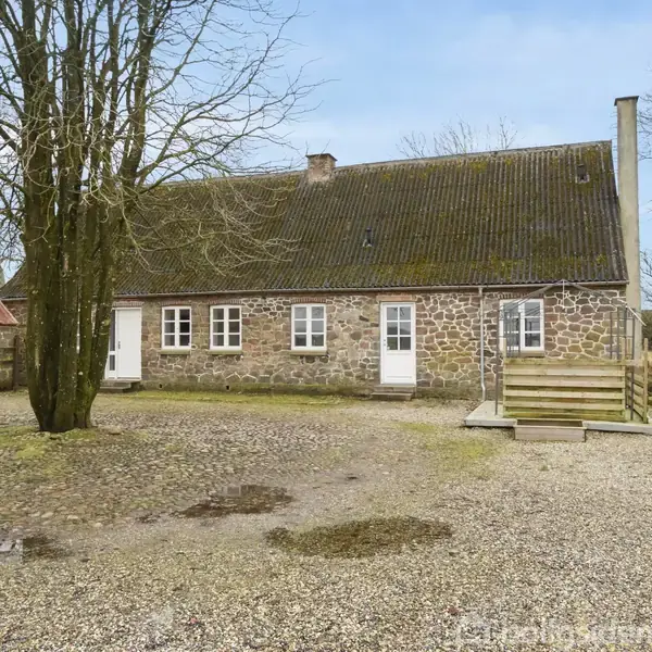 Et stenhus står stille på en bred gårdsplads med grus, omgivet af nøgne træer. Til venstre ses et rødt tag fra en anden bygning.