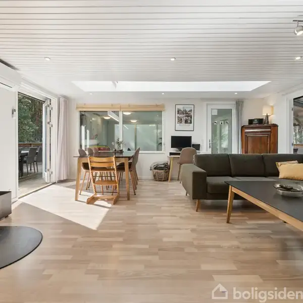 En moderne stue med brændeovn står til venstre, en grøn hjørnesofa med puder, et stort spisebord med stole midt i rummet, og glasdøre der åbner mod en terrasse udenfor.