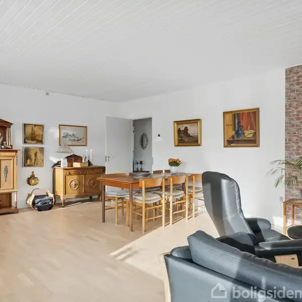 Stue med spisebord midt i rummet, omgivet af træmøbler og malerier på væggen. En lænestol står tæt ved en murstensvæg. Lys kommer ind fra et vindue uden for billedet.