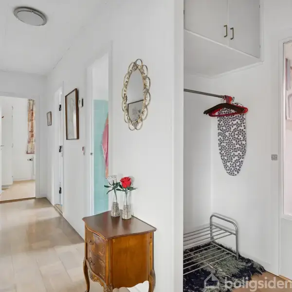 Et møbelbord med en vase med roser står i en lys gang. Væggen har et spejl, og der hænger strygebræt i et åbent skab. Døre fører til andre rum.