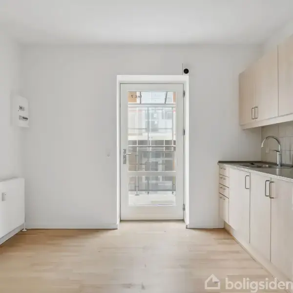 Et lyst køkken med trægulv. Skabene langs væggen er lyse, og der er en ovn og kogeplade. En dør i baggrunden fører ud til en balkon.