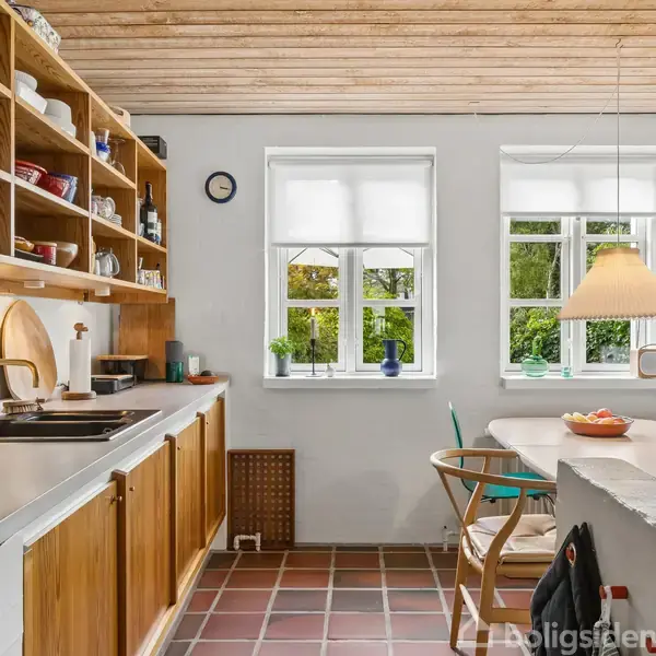 Køkken med trækøkkenelementer og åbne hylder fyldt med porcelæn; en vask med guldhane nær vinduer, der lukker naturligt lys ind; en bordplade med stole og en hængende lampe.