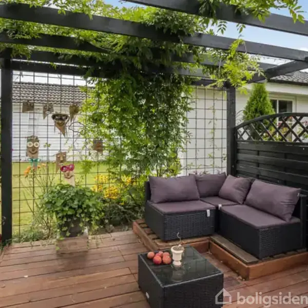 Sort loungesæt med lilla puder står på træterrasse omkranset af sort pergola med klatreplanter. En lille have ses i baggrunden med blomster og græsplæne.