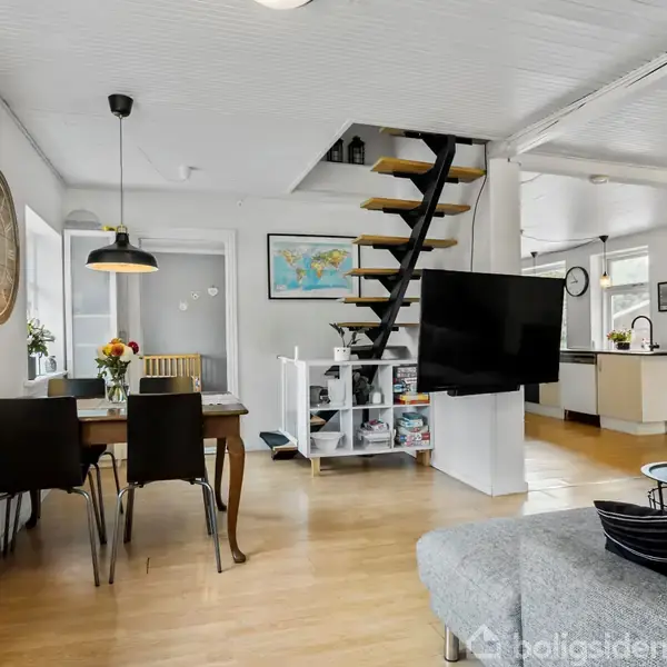 Et lyst stuerum med trægulv, spisebord til fire personer med blomster og en pendellampe, en trappe fører op fra sofaområdet, og et åbent køkken ses i baggrunden.