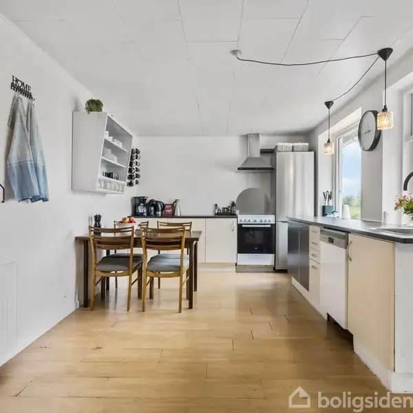 Køkkenbord med opvaskemaskine og vask står ved vinduer. Spisebord med tre stole i baggrunden. Hvide vægge med hylder, billeder og en opslagstavle fuldender rummet.