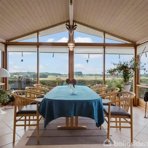 Et spisebord med otte stole er dækket med en blå dug og blomster. Rummet har store vinduer med udsigt over en frodig, landlig dal.