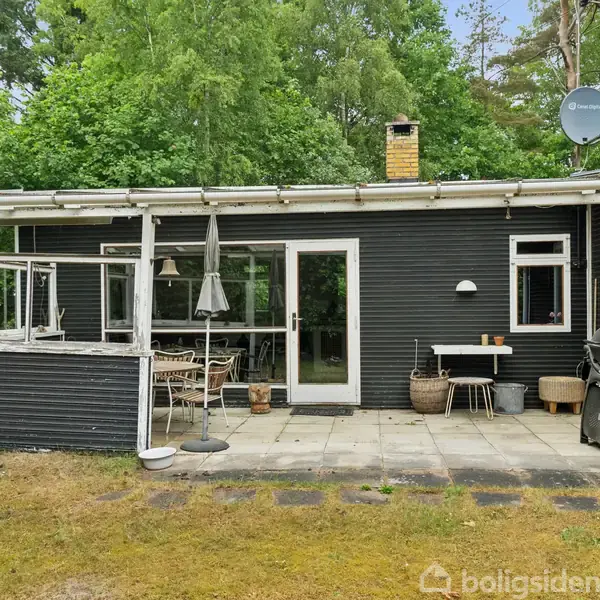 Sort træhus med hvide vinduesskodder står stille i en have omkranset af grønne træer. En sti fører til fordøren. Der findes ingen tekst i billedet.