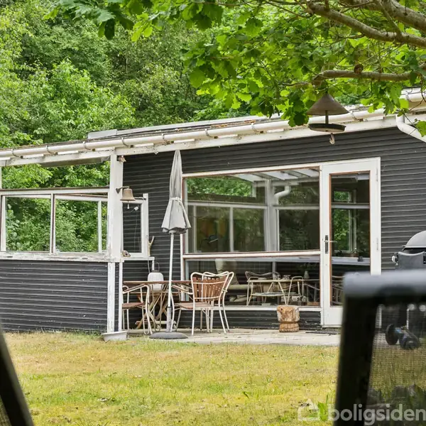 Et sort træhus med store vinduer står i en grøn have. Et sæt havemøbler og en parasol ses på terrassen, omgivet af frodig bevoksning.