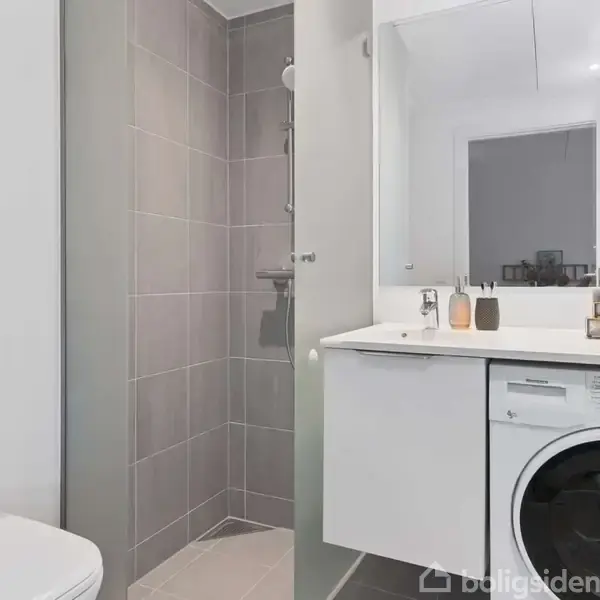 Badeværelse med vaskemaskine under en bordplade; bruser i glasbur til venstre; ovenfor et spejl og toalettsager. Minimalistisk og moderne indretning med grå fliser og lyse farver.