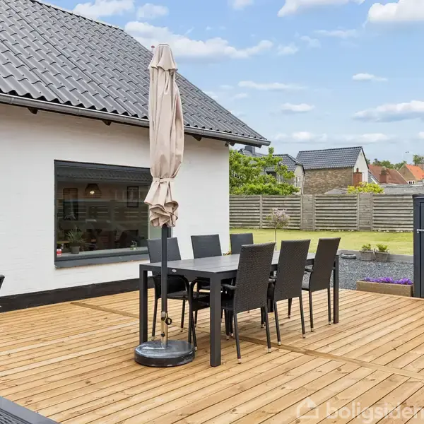 Terrasse med flisebelagt gulv. Møbleret med et bord, stole og en parasol. En terrassevarmer står ved siden af huset med en hvid væg, omgivet af et hegn og have.