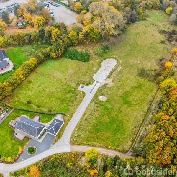 Grønne marker omgiver en vej og en bygning. To huse ligger i nederste venstre hjørne. Træer med efterårsfarver kantner billedet. парковка er synlig i øverste venstre hjørne.