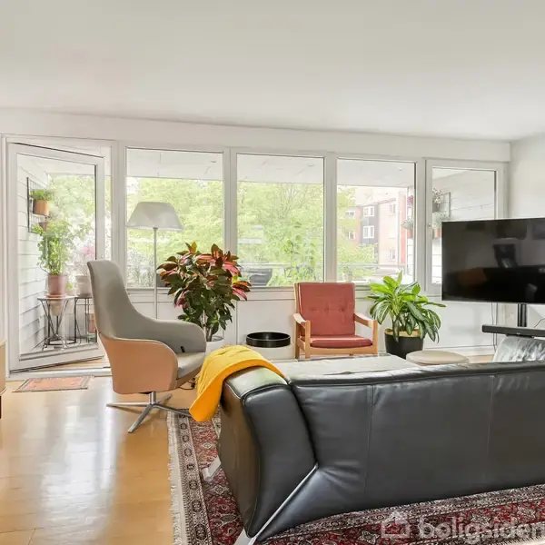 Sofa står centralt placeret, foran et stort fladskærms-tv monteret på væggen. Omgivet af lænestole, planter og et sofabord i en moderne, lys stue med vinduer langs væggen.