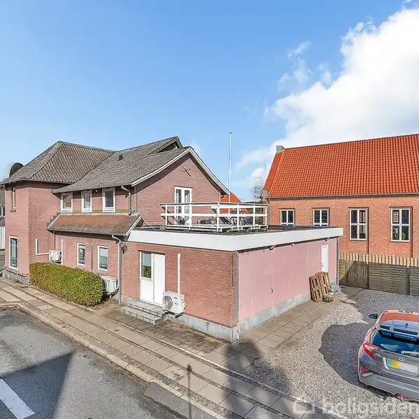 Et rødt murstenshus står ved en vej, flankeret af en parkeret bil til højre. Andre huse med røde tage ses i baggrunden under en blå himmel med skyer.