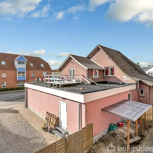 En pink bygning med fladt tag står ved en vej, omgivet af andre huse og bygninger. Skraldespande og en lille indhegning ses i forgrunden.