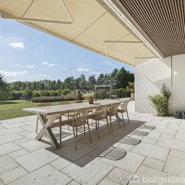 En lang træterrasse er udstyret med et stort bord og syv stole. Den ligger under en markise og vender ud mod en velplejet have med grønne planter og en græsplæne.