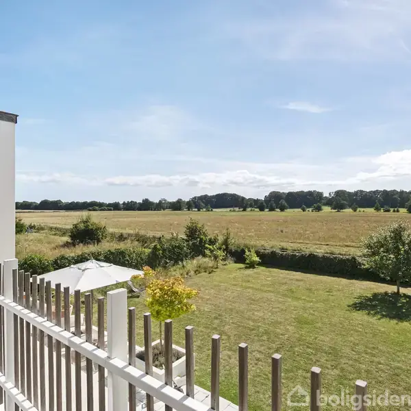 Balkon med trærækværk strækker sig ud mod en stor, åben mark under en blå himmel. Græsplæne og buske omgiver området, og en parasol ses nær et hus.