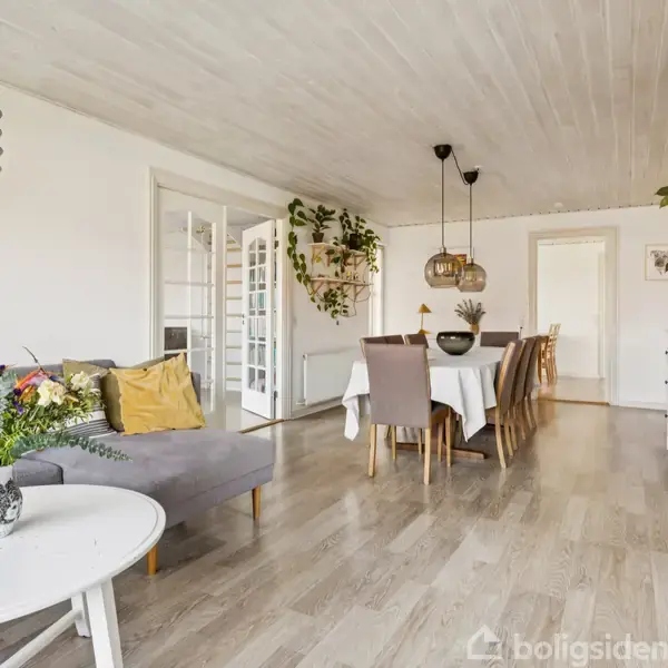 Sofa med farverige puder står ved lavt rundt bord med blomster, i rummelig stue med træloft. Spisebord med stole bagved, omgivet af grønne planter.