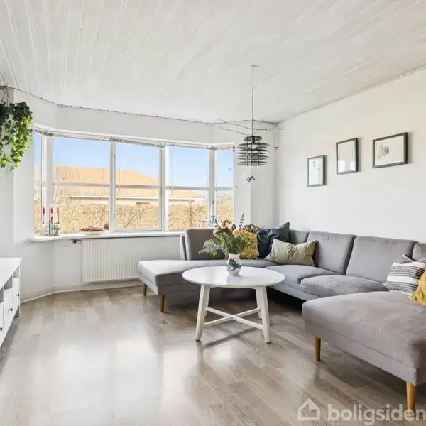 Stue med grå hjørnesofa omkring et rundt hvidt bord. Der hænger planter og lamper fra loftet. Store vinduer lader naturligt lys ind, og væggene har enkle dekorationer.