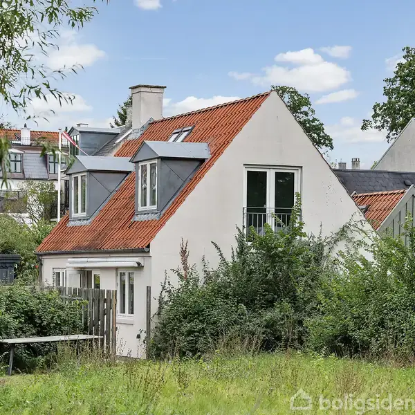 Hvidt hus med rødt tegltag og kvistvinduer står på en grøn græsplæne omgivet af buske og træer i et villakvarter. Himlen er blå med spredte skyer.
