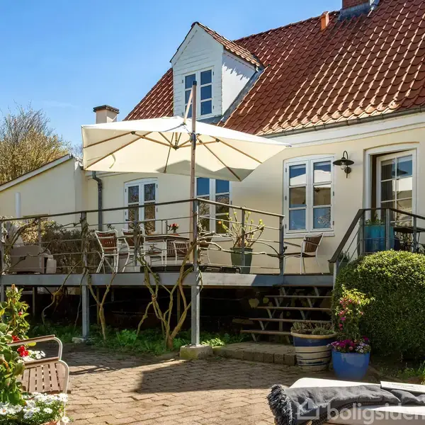 Et hvidt hus med rødt tag har en terrasse, der er udstyret med et stort parasol. Terrassen er omgivet af en have med farverige blomster og buske.