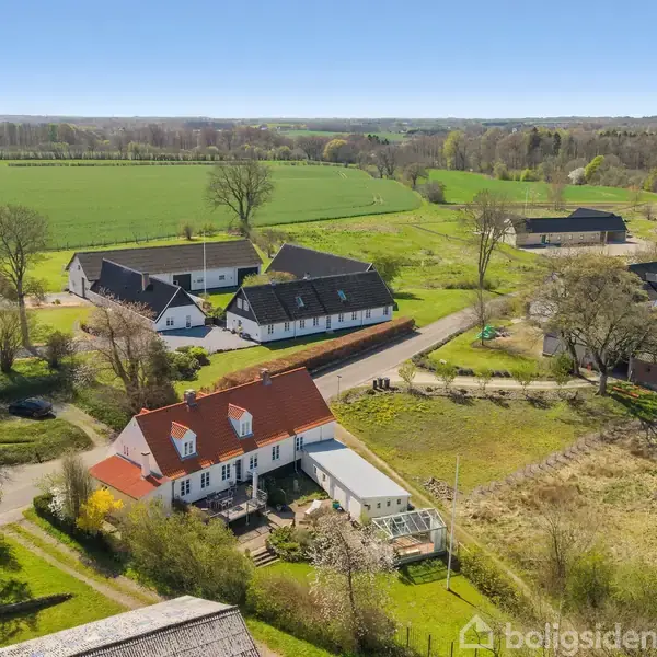 En hvid bygning med rødt tag står i et landligt område. Omkring huset er der grønt græs og træer. Flere bygninger og marker er i baggrunden under en klar blå himmel.