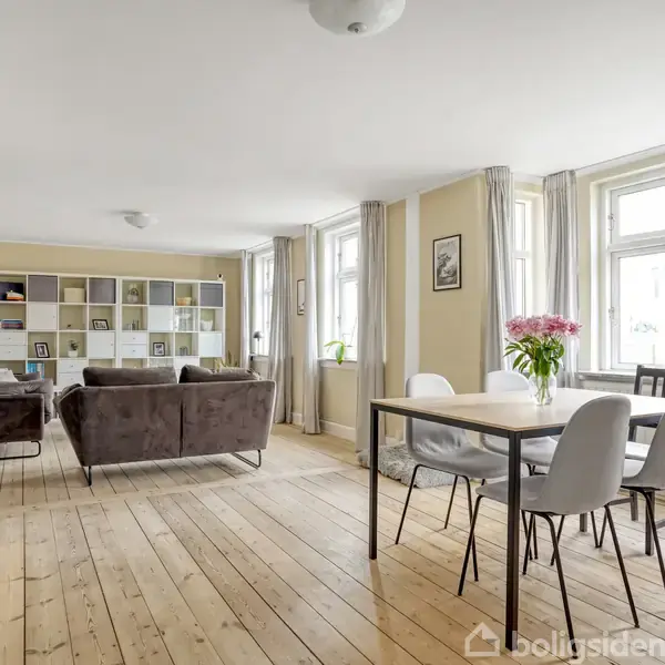 Stue med spisebord, fire stole og blomster. Sofaer og reoler i baggrunden. Lyse vægge og trægulv. Flere vinduer med gardiner slipper lys ind.