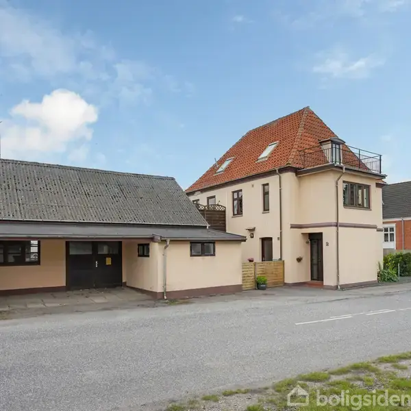 Bygning udført i lyse mursten med rødt tag, står ved en vej. To etager med vinduer, balkon. Ved siden er der en garage. Klart vejr med blå himmel og skyer.