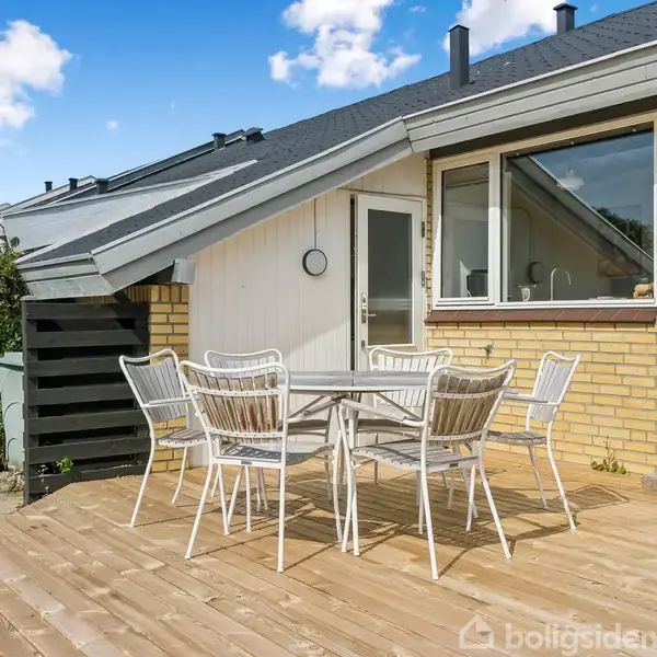 Terrassebord med seks stole står på træterrasse foran gul murstenshus. Bagved er trapper, en dør, et stort vindue og en klippet hæk. Himlen er blå med skyer.