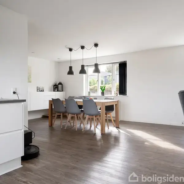 Et stilrent køkkenområde med et træbord omkranset af grå stofstole. Tre hængende lamper over bordet. Store vinduer lukker naturligt lys ind i det lyse rum.