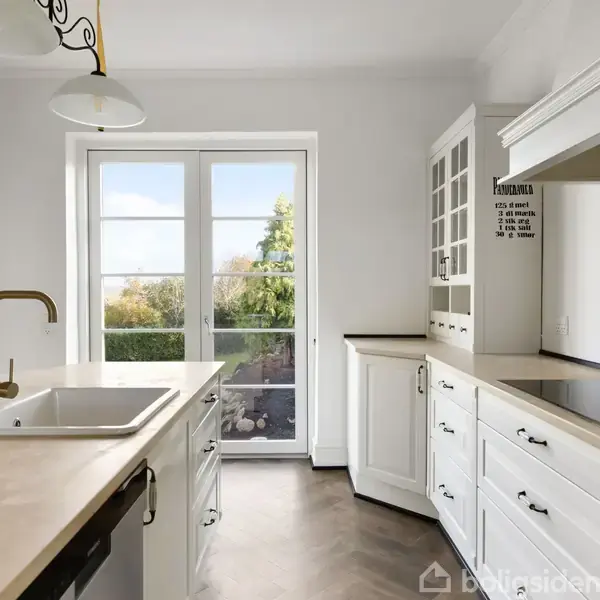 Et lyst og rummeligt køkken med hvidt inventar, vask og ovn i øen. Gennem store vinduer ses en have. På væggen står teksten: "KYLLINGER ER SÅ SMÅ AT DE NEMT KAN DØ SÅ HVIS DE ER SYGE SKAL DE VÆRE INDE".