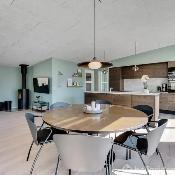 Køkken med moderne design har træskabe og central ø med vask. Sollys strømmer ind gennem store vinduer med persienner. I baggrunden ses en spisestue og udgang til terrasse.