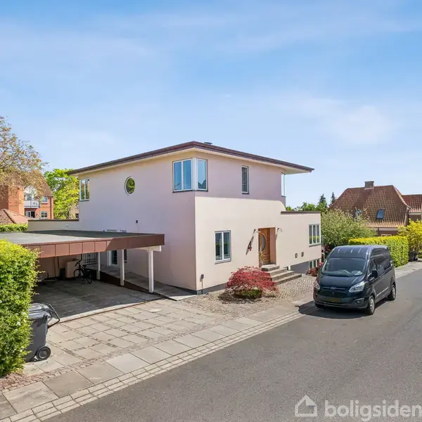 Et moderne to-etagers hus står på en stille villavej. En bil holder parkeret foran huset, som har en carport ved siden af og er omgivet af grønne buske og træer.