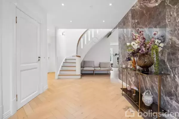 En elegant entré med en trappe, der svinger opad. Til højre står et bord med en vase fuld af blomster foran en marmoreret væg. Trægulv omgiver området.