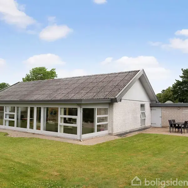 En hvid murstensbungalow med store vinduer står i en have med græsplæne. Til højre står et udendørs bord og stole. Himlen er blå med skyer.