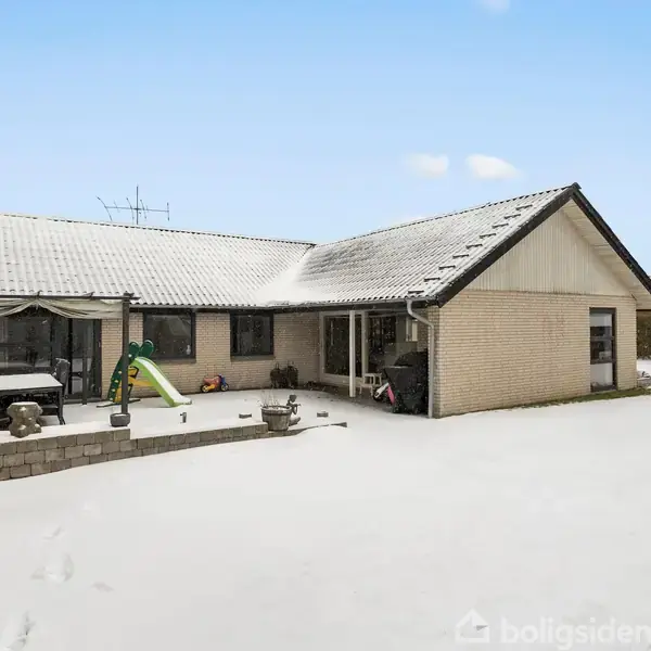 Hus med sneklædt tag står i en snedækket have. En terrasse med borde og stole er ved siden af, og et legehus med rutsjebane ses i baggrunden.