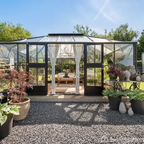 Drivhus med sorte rammer står i en have, omgivet af potteplanter og græsplæne. Indenfor ses et bord med blomsterdekorationer. Det er solskinsvejr, og himlen er blå.