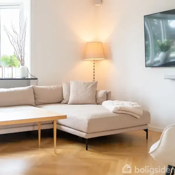 En lys stue med en beige hjørnesofa, bord, og sidebordslampe. Fjernsyn hænger på væggen. Store vinduer giver udsigt til grønne omgivelser. Ingen tekst til stede.