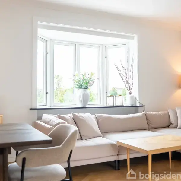 En hvid sofa står i hjørnet af en lys stue. Ved siden af sofaen er et træbord. En stor vindueskarm indeholder planter. En lampe lyser i hjørnet.