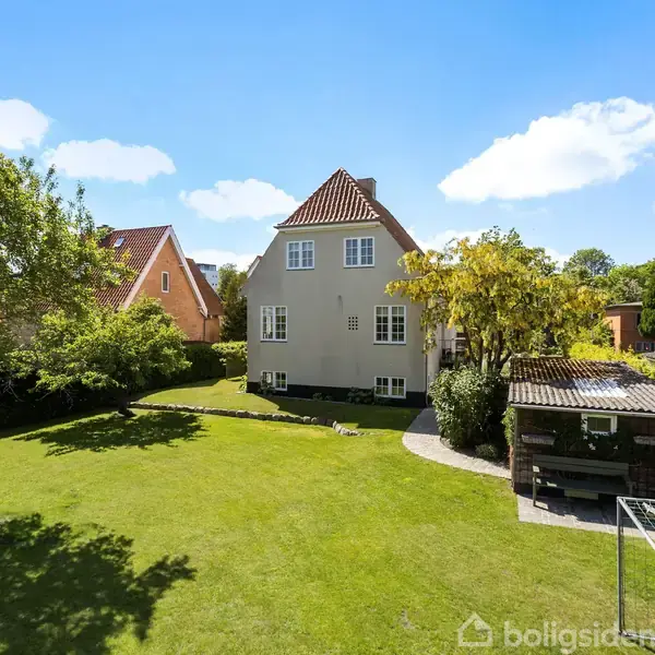 Et hvidt hus med rødt tag står i en frodig have. Solen skinner, og himlen er blå med få skyer. Til højre ses et skur med overdækning.