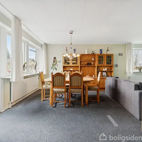 Spisebord med seks stole står i en stue med lyse vægge og store vinduer. Reol i baggrunden og gråt tæppe på gulvet.