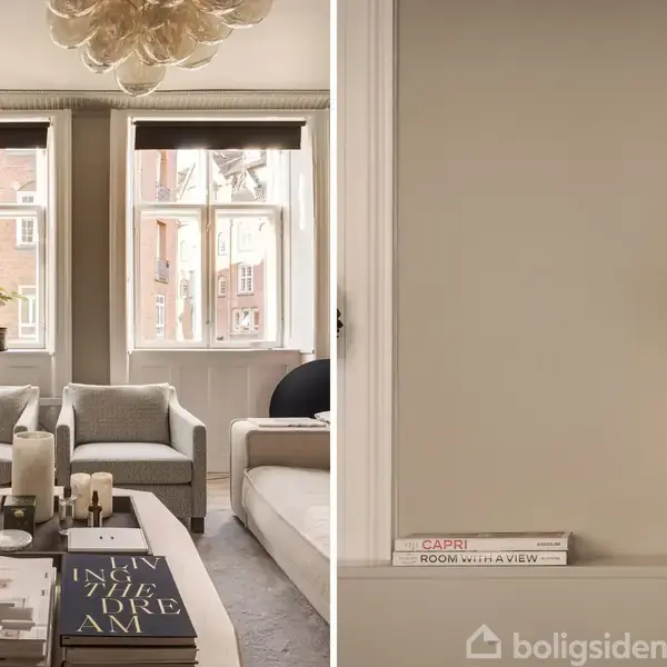 En hyggelig stue med sofagruppe og sofabord, pyntet med bøger og lys. To store vinduer med udsigt. På en hylde ses bøgerne "CAPRI" og "ROOM WITH A VIEW" ved siden af en lampe.